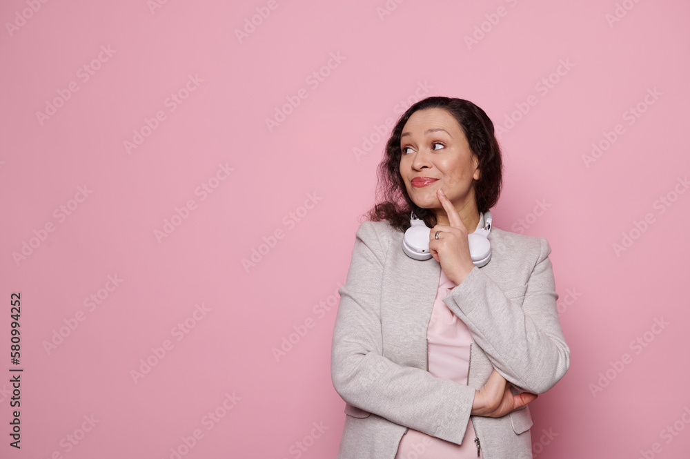 Pensive middle-aged pretty woman with wireless headphones, dreamily looking aside at copy space for advertising text, posing with arms crossed, holding her finger on chin over isolated pink background Pensive middle-aged pretty woman with wireless headphones, dreamily looking aside at copy space for advertising text, posing with arms crossed, holding her finger on chin over isolated pink background