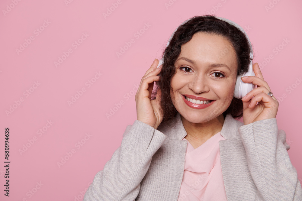Headshot of a multi-ethnic middle-aged woman wearing wireless headphones, a music lover, enjoying new playlist and sound track, smiling with a beautiful toothy smile looking at camera, pink background Headshot of a multi-ethnic middle-aged woman wearing wireless headphones, a music lover, enjoying new playlist and sound track, smiling with a beautiful toothy smile looking at camera, pink background