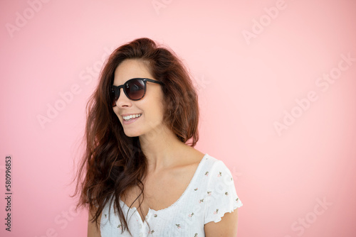 pretty young woman with black sunglasses and long hair with flowered shirt is standing in front of pink background © epiximages