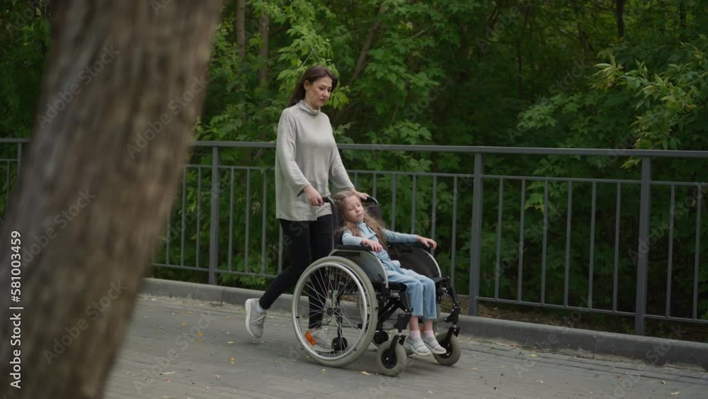 Mom walks on pavement with little daughter sleeping in wheelchair. Adult woman takes girl with muscular dystrophy out to city park on spring day Mom walks on pavement with little daughter sleeping in wheelchair. Adult woman takes girl with muscular dystrophy out to city park on spring day