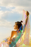Young woman posing with holographic foil against sky. Dreamy self expression concept fashion portrait