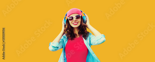 Colorful portrait of happy smiling young woman listening to music in headphones wearing pink hat on orange background © rohappy