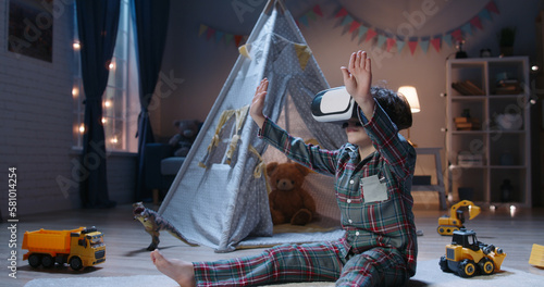 Copy space. Little boy is playing video games before bedtime, using virtual reality headset and experiencing new world. The future is the present. Happy smart kid with curly hair with VR headset 