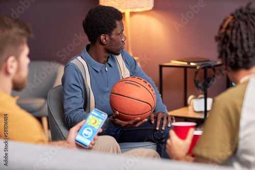 Side view at diverse group of sports fans watching basketball match at home and using online betting app