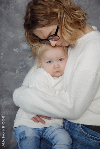 Young mother, dressed in jeans and white sweater gently hugs her little daughter dressed just like her. Motherhood. 