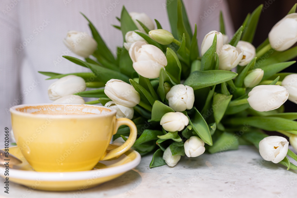 Yellow ceramic mug with cappuccino is on table in cafe, next to it lies luxurious bouquet of tulips. Floral surprises. Yellow ceramic mug with cappuccino is on table in cafe, next to it lies luxurious bouquet of tulips. Floral surprises.