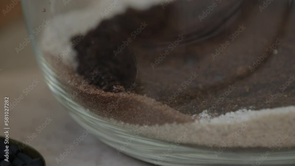 A young female botanist creates a tiny live forest ecosystem in a glass terrarium - putting brown sand layer - a tight close-up A young female botanist creates a tiny live forest ecosystem in a glass terrarium - putting brown sand layer - a tight close-up