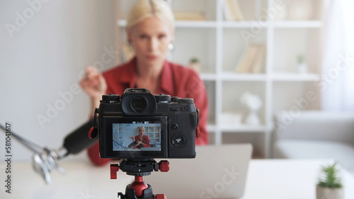 Influencer vlog. Live streaming. Online coaching. Mature blogger woman talking recording video interview on camera on tripod on defocused interior with free space.