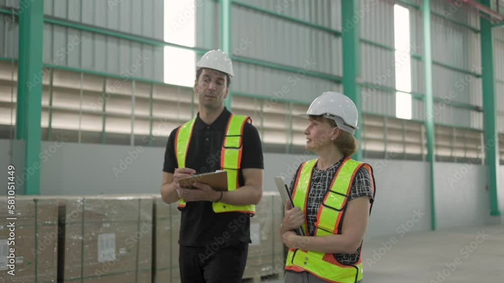 Caucasian senior female manager wearing safety vest holding laptop and male engineer walking warehouse inspection Caucasian senior female manager wearing safety vest holding laptop and male engineer walking warehouse inspection