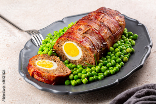 Baked meatloaf made of ground meat, onion, carrot, stuffed with hard-boiled eggs, wrapped with bacon net, served with green peas. German, Scandinavian and Belgian dish. Festive and Easter food. photo
