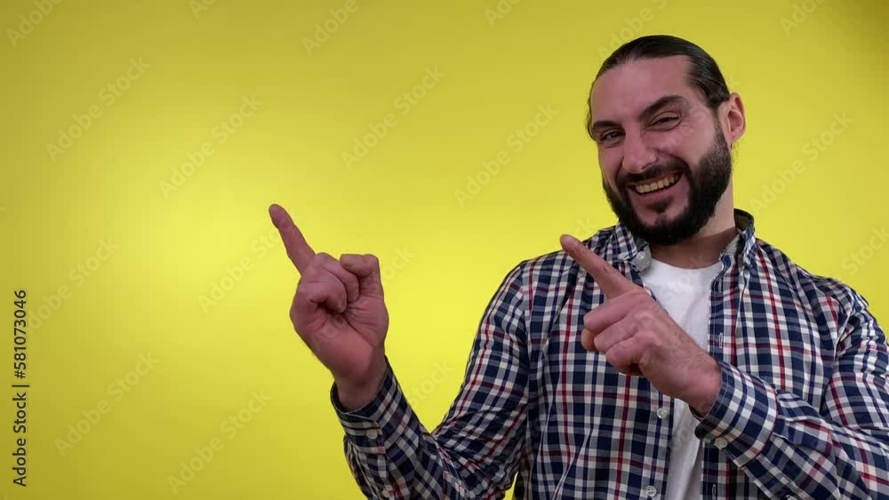 Positive bearded man pointing fingers away paying attention at empty space for advertisement, looking at camera with toothy smile isolated on yellow background. Announcement Advertisement Concept Positive bearded man pointing fingers away paying attention at empty space for advertisement, looking at camera with toothy smile isolated on yellow background. Announcement Advertisement Concept