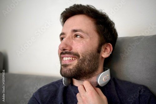 Young man wearing electric pulse neck massager while sitting on the sofa 