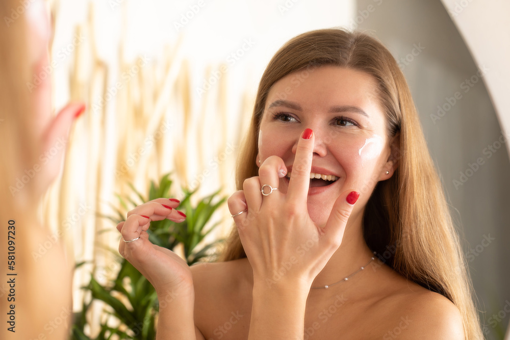 A joyful woman puts a moisturizing anti-aging cream on her face in front of the bathroom mirror A joyful woman puts a moisturizing anti-aging cream on her face in front of the bathroom mirror