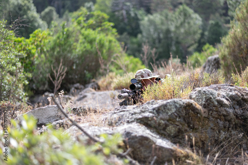 Homme militaire en uniforme en position de tir entre rochers de granit et végétation
