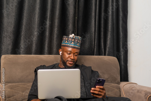 Good looking businessman in Africa attire seated using his laptop and using his mobile phone to perform business transactions