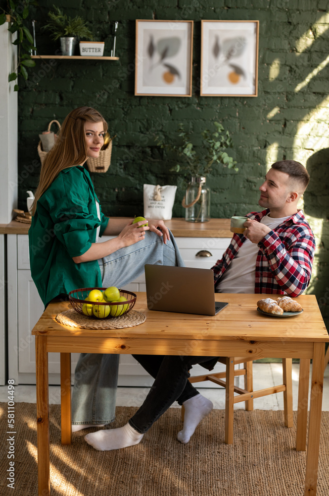 Cute couple in the kitchen. A man with a cup of coffee in his hand is sitting in front of a laptop looking at his wife Cute couple in the kitchen. A man with a cup of coffee in his hand is sitting in front of a laptop looking at his wife