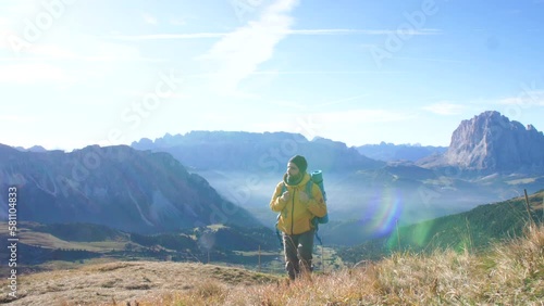 Young man hiking at Seceda mountain arriving to peak at sunrise. Backpack, yellow jacket, boots, beanie. Traveling to puez Odle, Dolomites, Trentino, Italy.