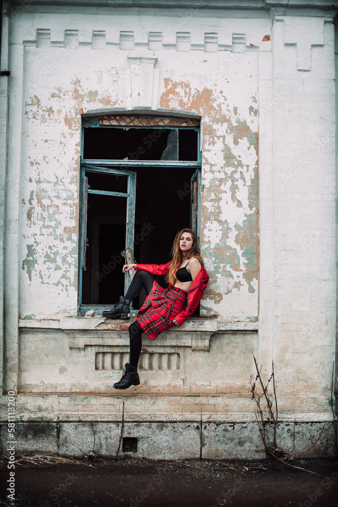 A sad young girl in a red jacket, bra and red plaid skirt sits in the window frame of an old ruined building on a city street.