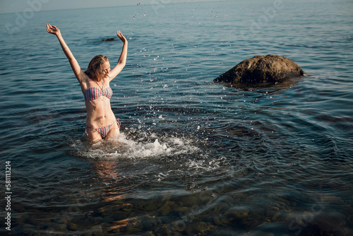 A blonde girl with long hair, in a striped swimsuit, bathes in the ocean, laughs, plays and splashes on a bright sunny day.