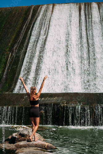 A slender tanned girl with a sports figure is doing yoga exercises against the backdrop of a picturesque waterfall in the summer. She is wearing black shorts and a top.
