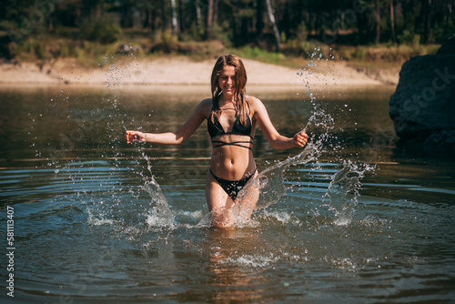 A young girl with long blond hair, a slender figure, in a black swimsuit jumps in the river against the backdrop of rocks and laughs.