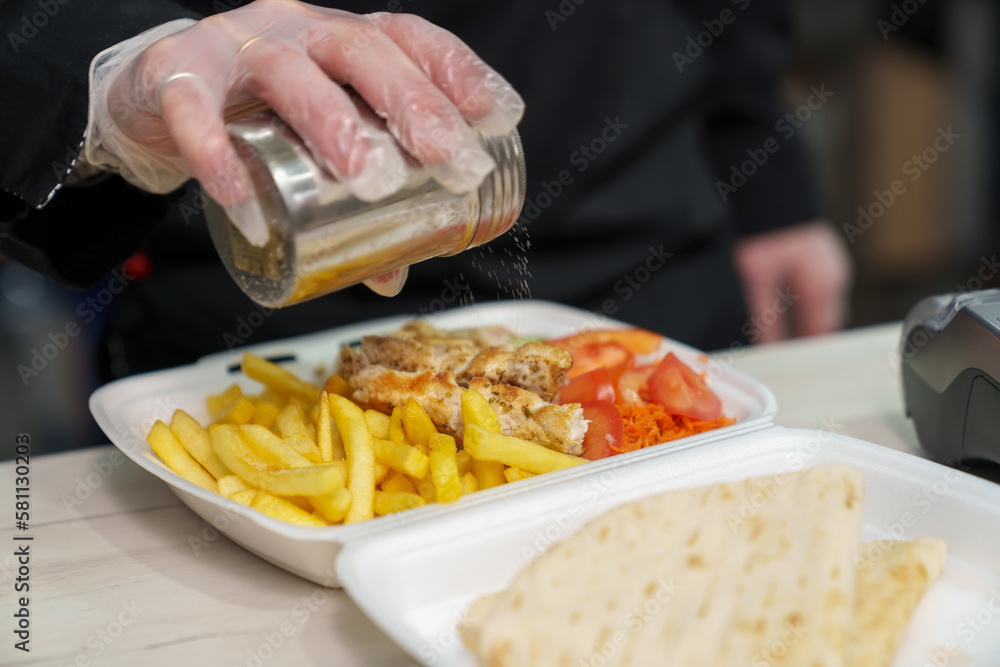 Cook seasoning lunch box with marinated souvlaki meat and fries with salt. Chef cooking traditional Greek fast food