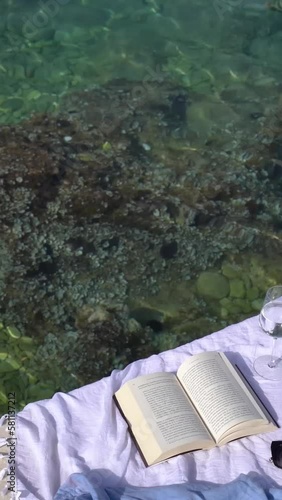 A vertical video of a book on a white beach blanket with a blue shirt and a glass of wine next to the sea