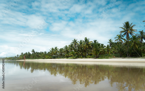 Pessoa andando em praia paradisíaca rodeada de coqueiros e com reflexo na água (ID: 581142440)