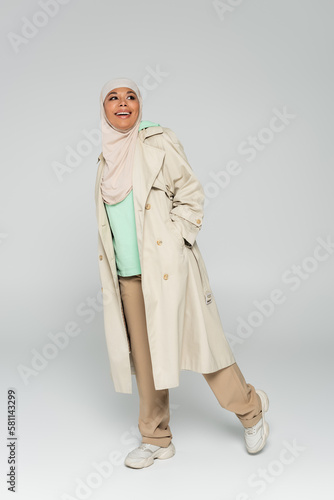 full length of multiracial muslim woman in hijab and trench coat with sneakers posing with hand in pocket on grey background.