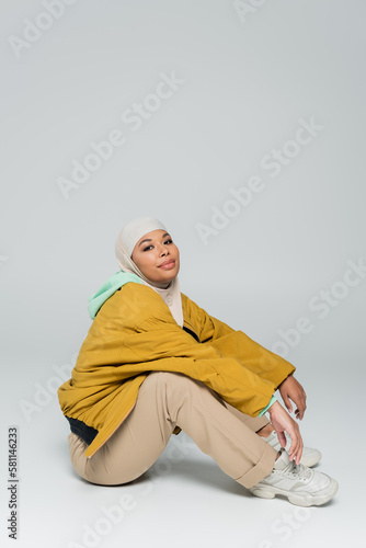 full length of pleased multiracial muslim woman sitting in beige pants and yellow jacket on grey background. © LIGHTFIELD STUDIOS