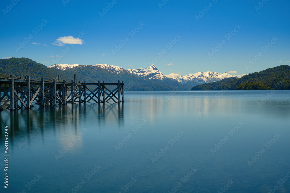 A beautiful pier to the left of the lake in shades of green and blue with a background of mountains in the morning.