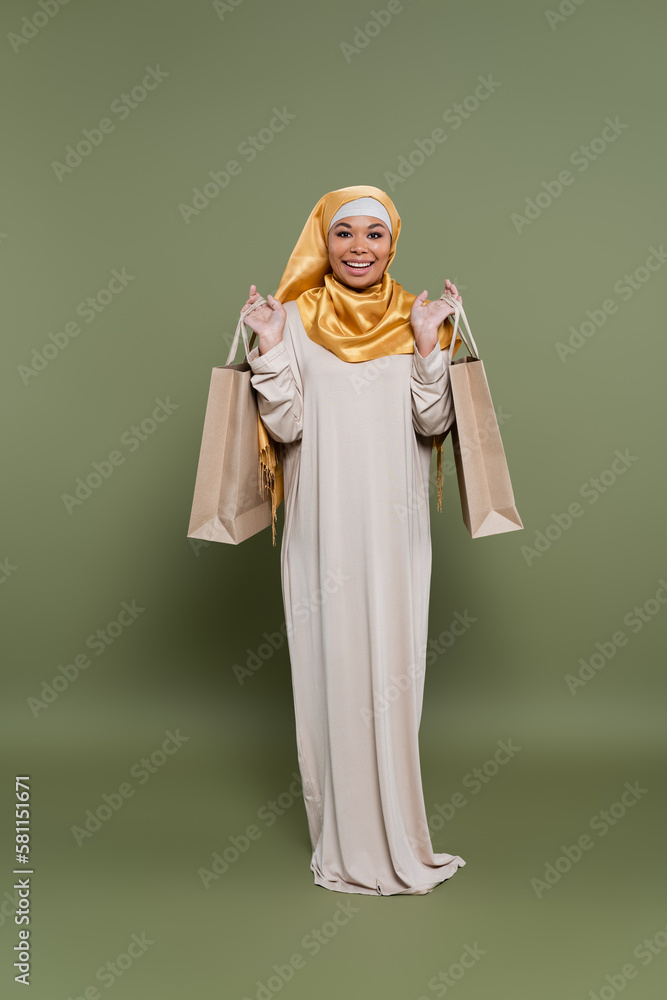 Positive multiracial woman in hijab holding shopping bags on green background.