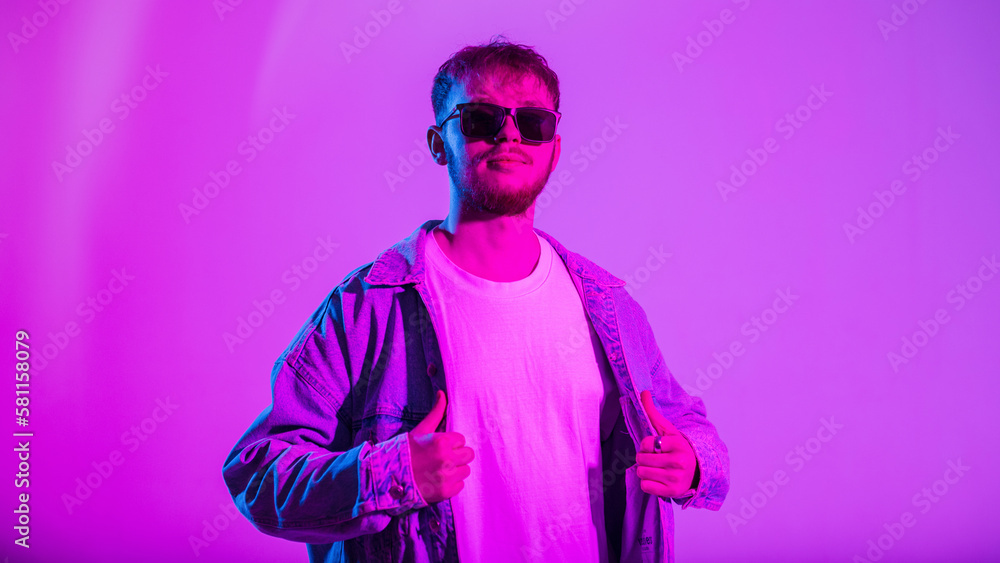 Stylish trendy handsome hipster guy with sunglasses in fashion denim clothes with jacket and t-shirt in color studio with creative pink and neon light