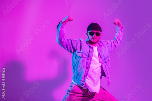Stylish handsome professional hip hop dancer man with cap and sunglasses in trendy fashion denim clothes dancing in studio with creative pink and neon light