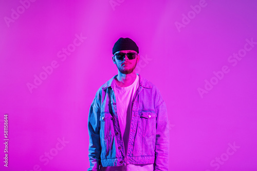 Stylish handsome creative young guy with a cap and sunglasses in a trendy fashion denim jacket in a studio with pink and neon lights