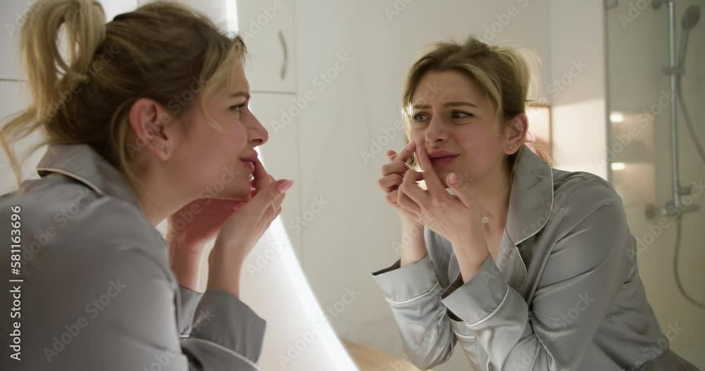 Young woman is looking at acne pimples on her face in the mirror in the bathroom. Problematic skin Young woman is looking at acne pimples on her face in the mirror in the bathroom. Problematic skin