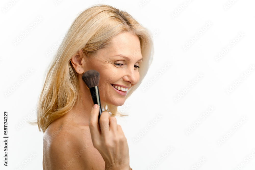 Beautiful 50s middle-aged woman holding makeup brush near chin and applying powder, eyes closed dreamly, charming lady isolated on white background. Skincare concept Beautiful 50s middle-aged woman holding makeup brush near chin and applying powder, eyes closed dreamly, charming lady isolated on white background. Skincare concept