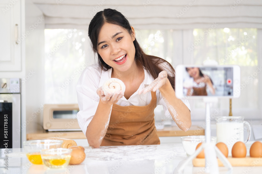 Portrait of smiling young woman food vlogger showing process of baking in live stream, standing in front of smart phone on tripod, broadcasting. Indoor studio shot on kitchen background.