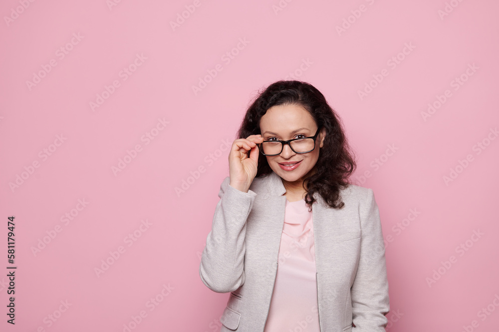 Beautiful dark-haired middle-aged woman in stylish elegant suit and spectacles, smiling looking at camera on isolated pink background. Eyesight. Health. Medicine. Ophthalmology and fashion concept