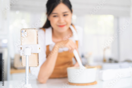 baker online courses. food preparation and culinary training class concept. smiling woman chef prepare ingredients in the kitchen and shooting video of herself using mobile phone on a tripod. photo