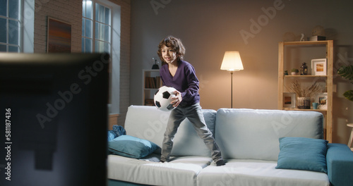 Funny expressive little asian boy watching a soccer game on tv and jumping on couch in front of it, reacting to his favourite team playing - football fans concept