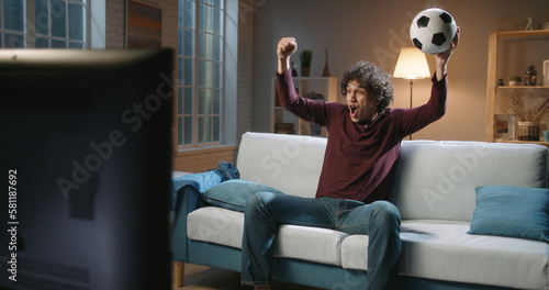 Expressive Spain man football team supporter emotionally watching a soccer game. Hipster guy with curly hair watching tv at home alone 