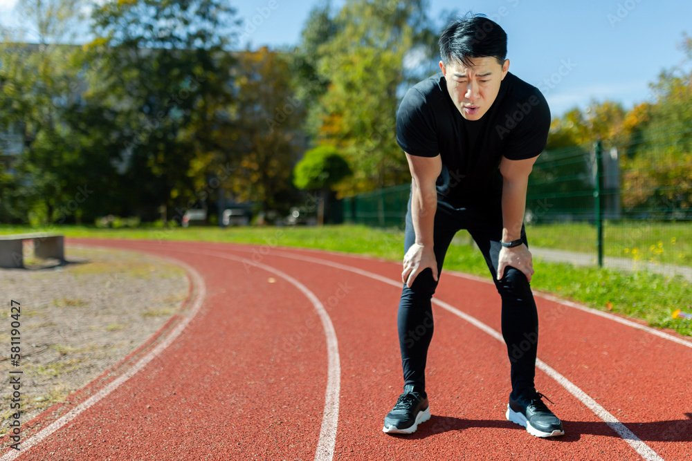 Obraz premium Asian sportsman tired after running and active physical exercise, man taking breath and rest on sunny day in stadium, overtired after running.