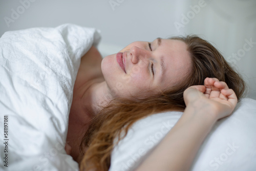 Young happy beautiful sleepy woman in the bed in bedroom at home in the morning lying under white blanket, enjoy resting, sleep well, wake up and smile. Good morning