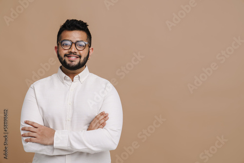 Young indian man standing with arms folded isolated over beige wall