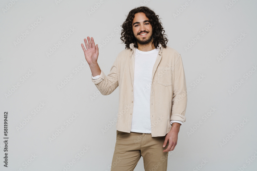 Smiling hispanic man hair waving hand while standing isolated over white wall Smiling hispanic man hair waving hand while standing isolated over white wall