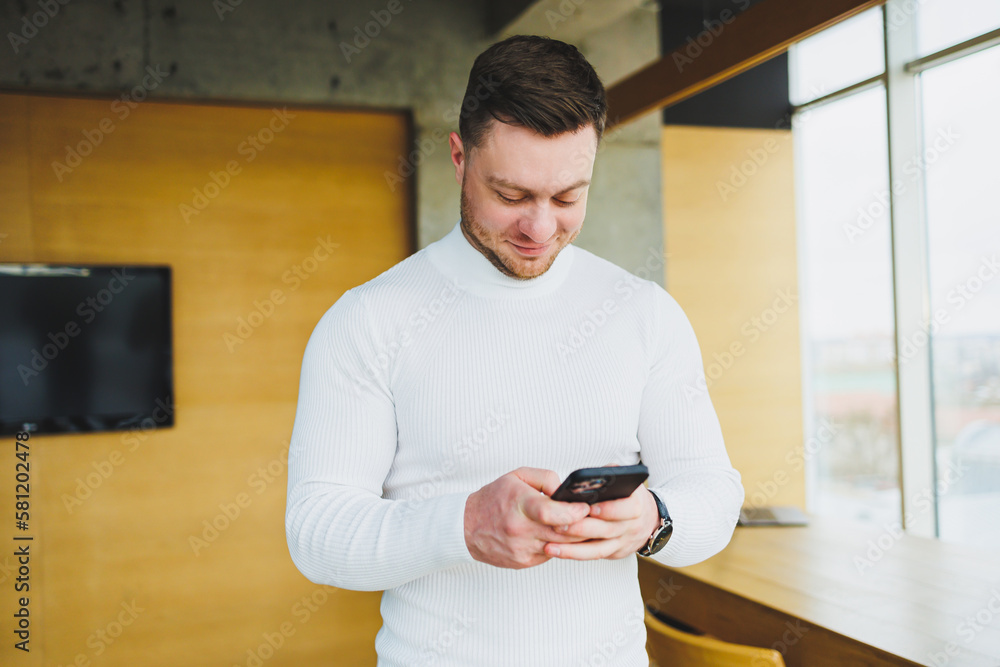 A young man in a white pullover is standing in a spacious office and talking on the phone. A man talks to colleagues via video link. A young man in a white pullover is standing in a spacious office and talking on the phone. A man talks to colleagues via video link.