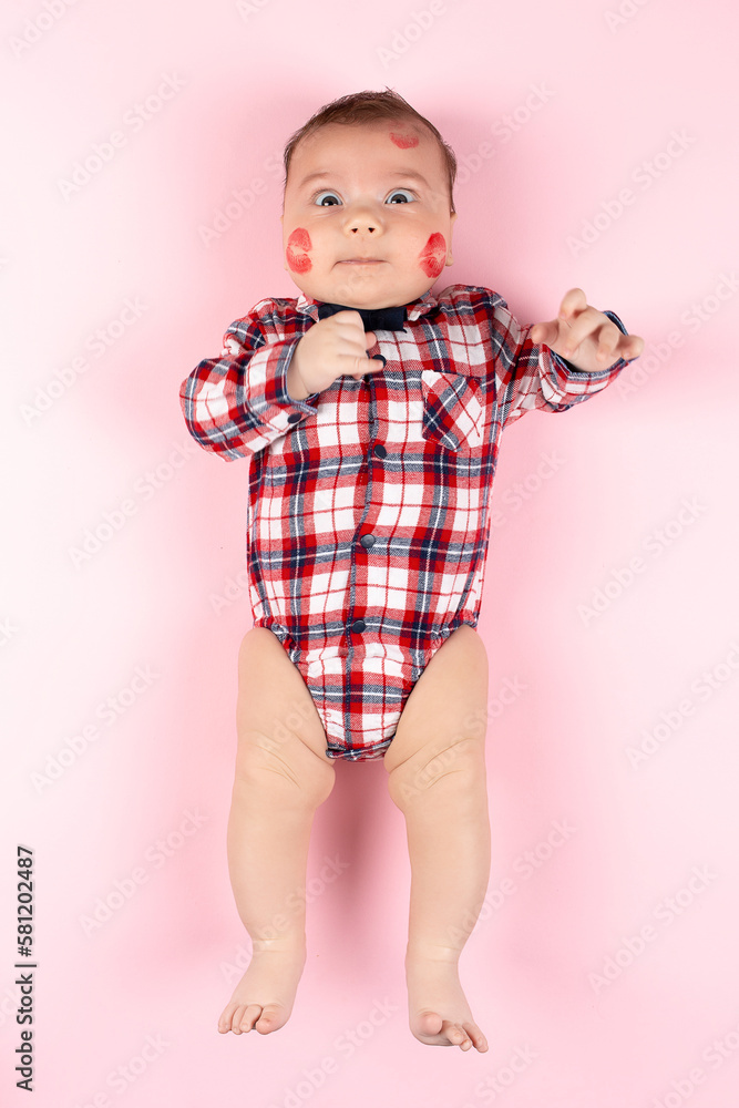 Child in beautiful plaid shirt on pink, traces of kisses with red lipstick on his face. Mockup for advertising, design, postcards, Valentine's Day. Child in beautiful plaid shirt on pink, traces of kisses with red lipstick on his face. Mockup for advertising, design, postcards, Valentine's Day.