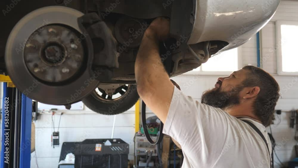 Repairman in uniform installs a new part to car at workshop. Worker servicing automobile to changing belt. Professional bearded auto mechanic works underneath a lifting vehicle at garage. Slow motion