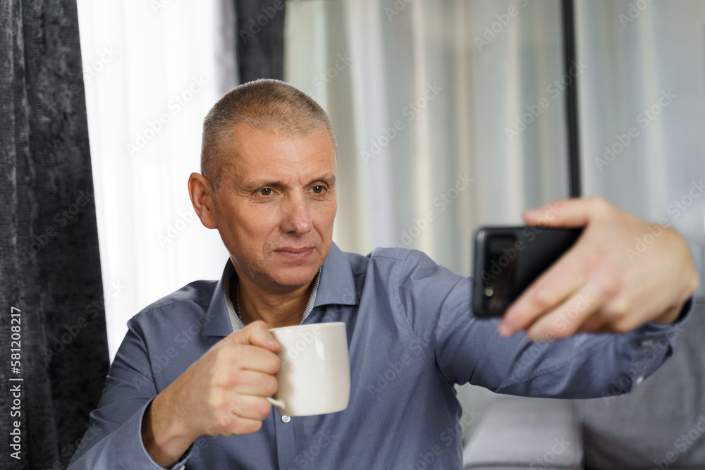 A man with tea makes a selfie photo with a mobile phone in the apartment. A man with tea makes a selfie photo with a mobile phone in the apartment.